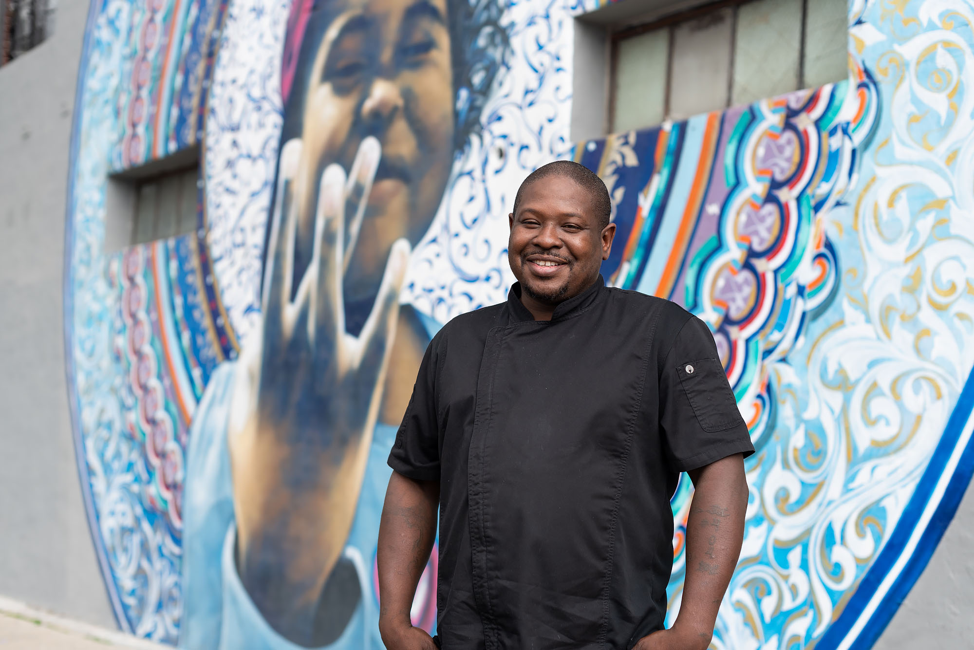 A portrait of a man, Keith Corbin, standing in front of a wall painted with a mural. 