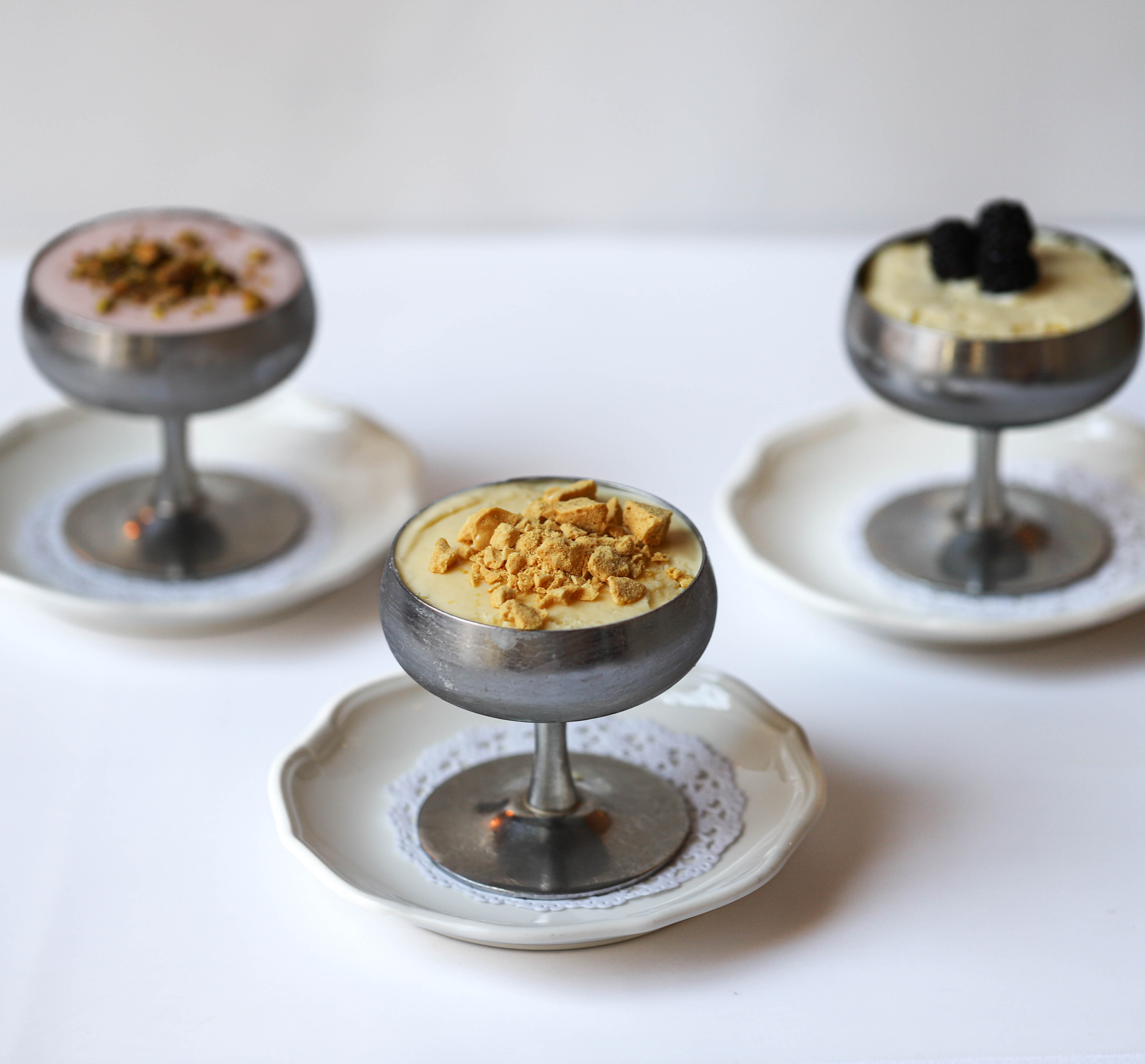 three metal coupe glasses of ice cream sit on top of doilies on classic ceramic plates on a white counter in front of a white background