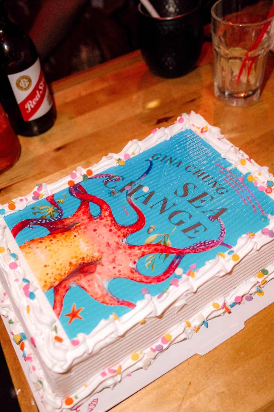 A sheet of Carvel ice cream cake decorated with the cover of Gina Chung’s book Sea Change. The cover features a large pink octopus with its tentacles outstretched in front of a turquoise background. Chung’s name and the book’s title are above it. The cake is piped around the edges with white frosting and dotted with colorful, round sprinkles.
