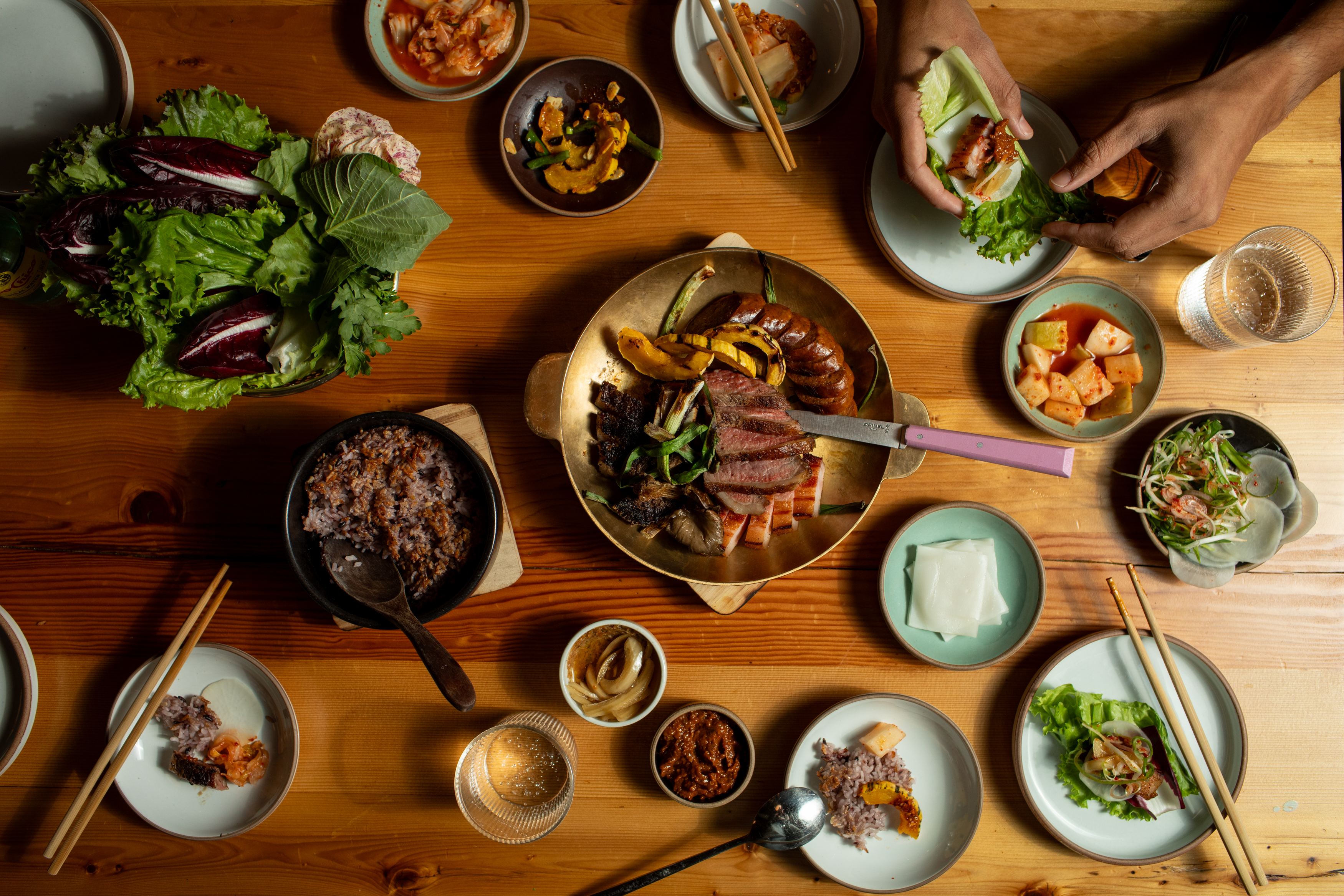 Overhead look at a table filled with cuts of meat, rice, and banchan. Two hands assemble a bite of meat and pickles on a lettuce wrap.