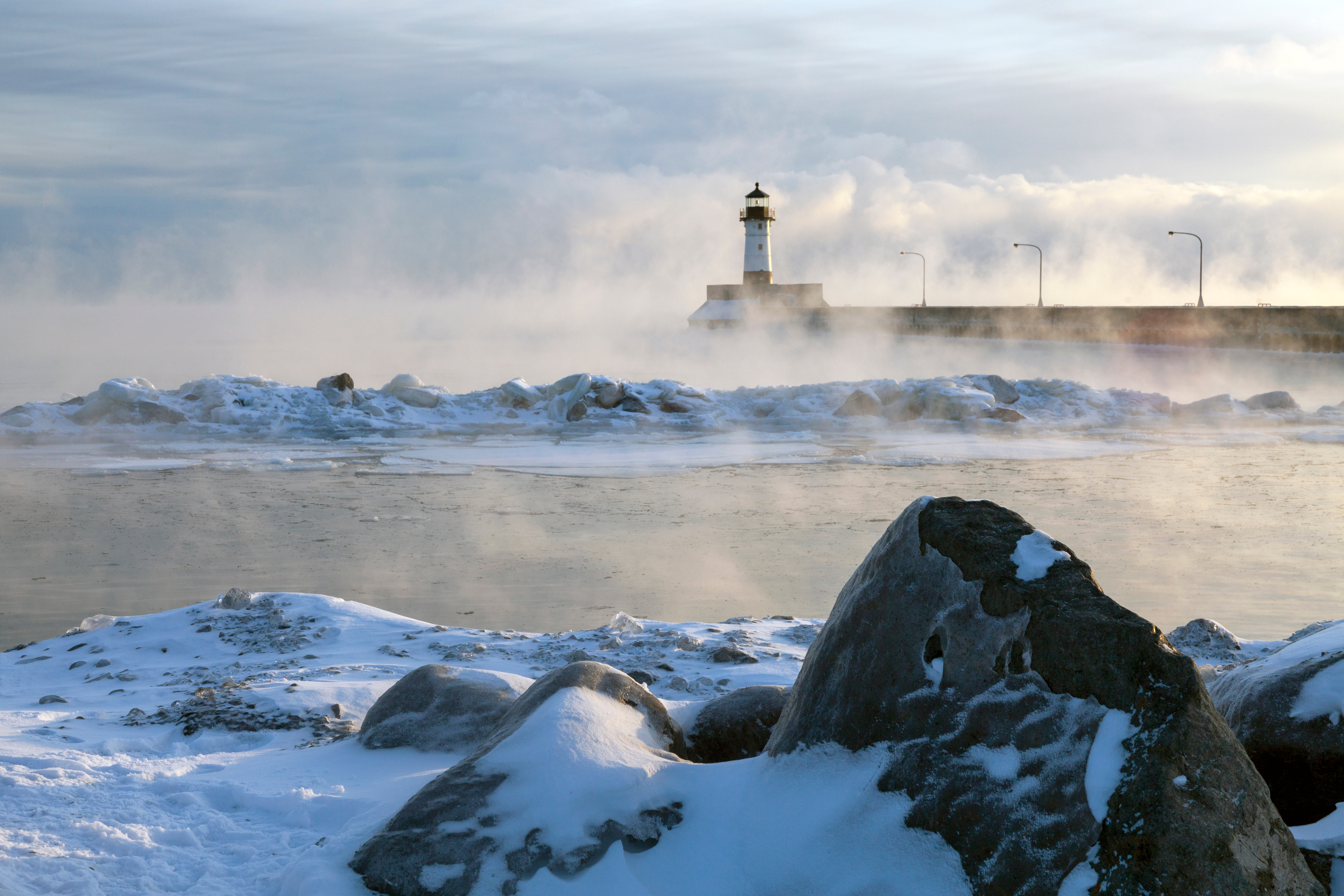 A view of a pier and lighthouse reaching out into Lake Superior in the winter, with mist rising from the water, and snow-covered rocks in the foreground. 